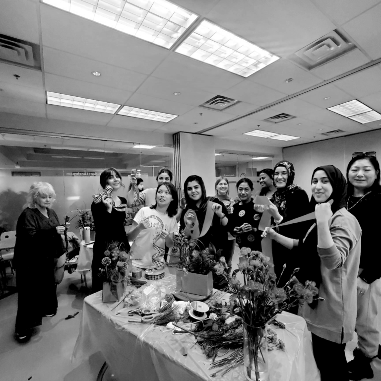 A cheerful group of women of diverse age and ethnicity are gathered around a table at an event, holding ribbons and roses