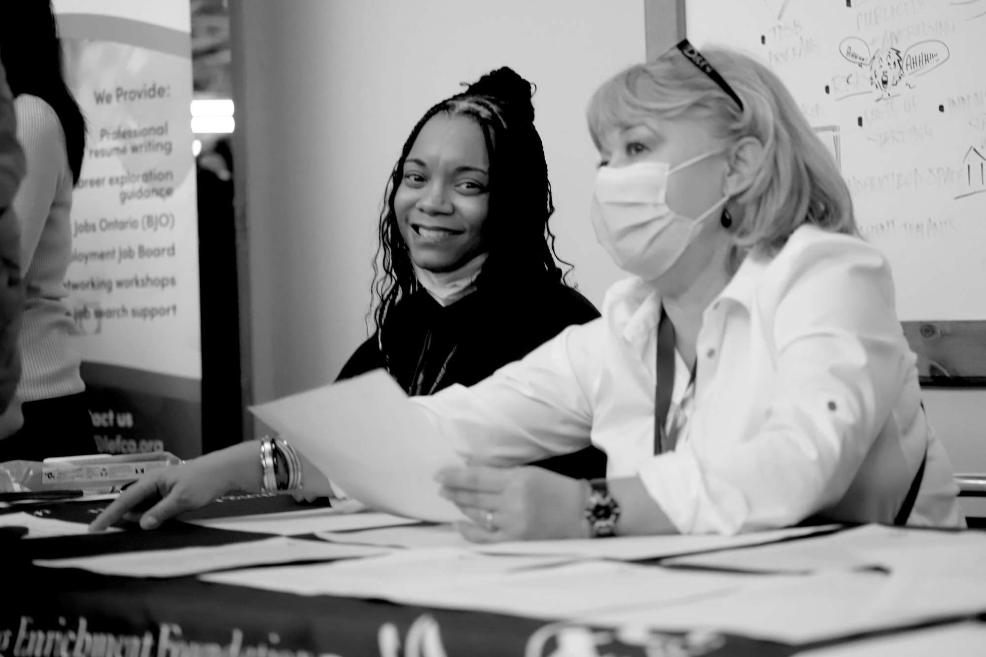 Two women sitting at a table in an event are interacting with event guests. One of them has a mask on her face. The other one has her mask pulled down and is smiling into the camera