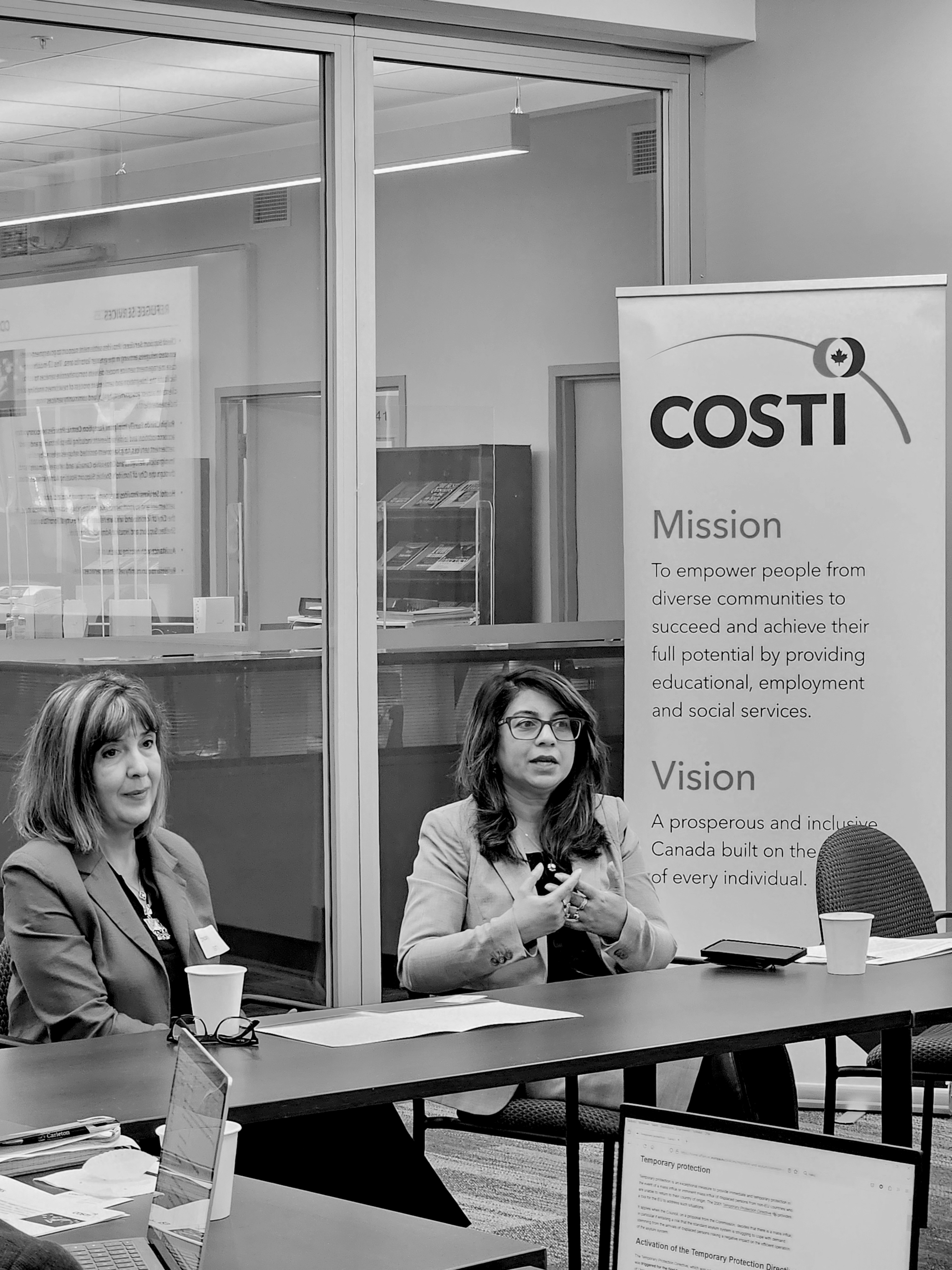 Director of Education, Snezana Gabric, and Interim Associate Director of Social Services, Rahila Mushtaq addressing an audience, sitting at a desk in front of COSTI standee