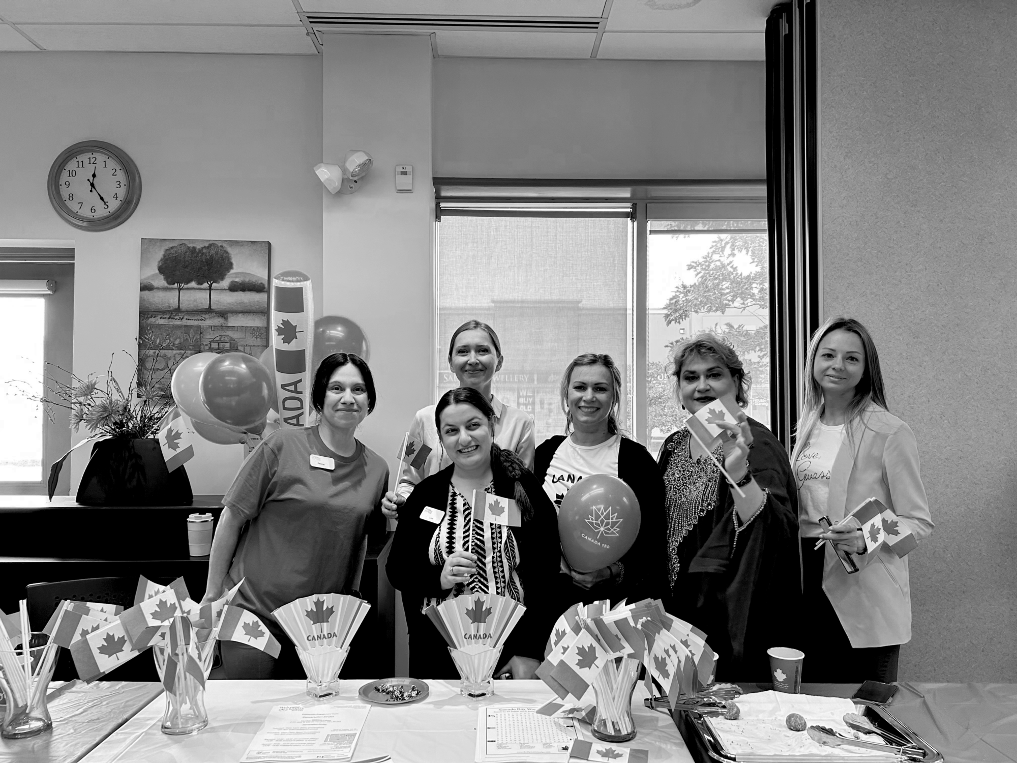 A diverse group of women standing together, each holding a small flag of Canada, are standing behind a desk full of Canadian flag merchandize, smiling for the camera