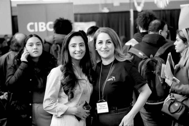 Two COSTI employees at a hiring event, with a crowd of prospective clients and partner organizations in the background.