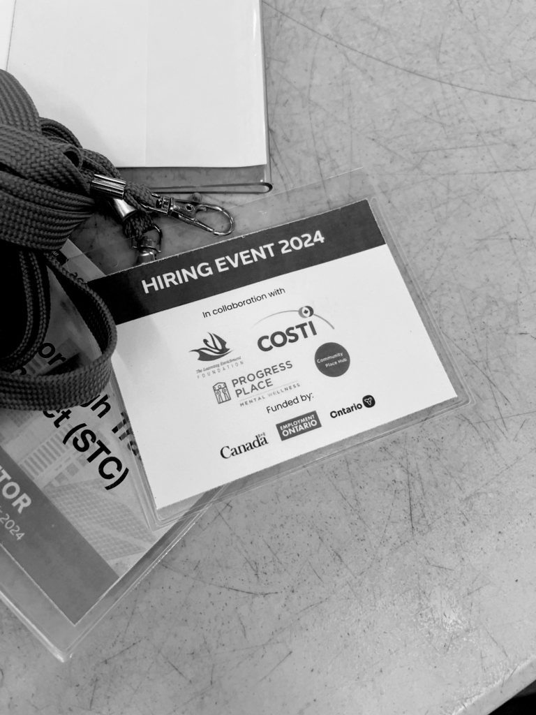 A white and teal coloured lanyard of COSTI hiring event with logos of funders and partners, kept on a table.