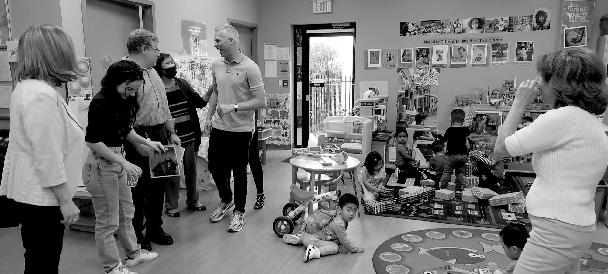 A group of individuals talking and laughing in the corner of a playgroup area, while some kids are playing in the background