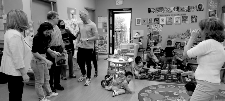 A group of individuals talking and laughing in the corner of a playgroup area, while some kids are playing in the background