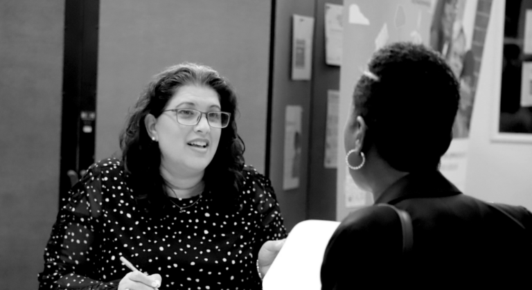 : Two women sitting on opposite sides of a table, talking to each other. One of them is taking notes with a pen and paper in hand