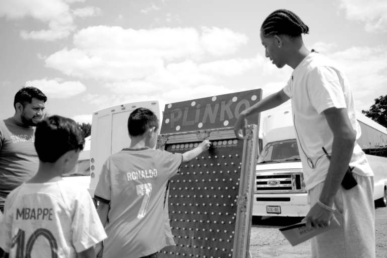 Two young boys are playing a board game called Plinko in an outdoor setup. One male adult is helping them, and another one is enjoying looking at the game.