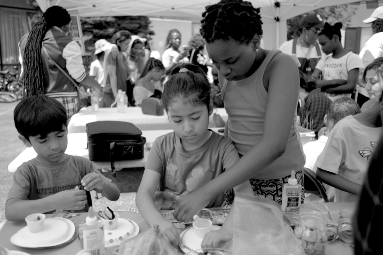 Two kids are engaged in art and craft work at a table in an outdoor setup, under the supervision of a young adult. In the background, many more children and supervisors are doing the same