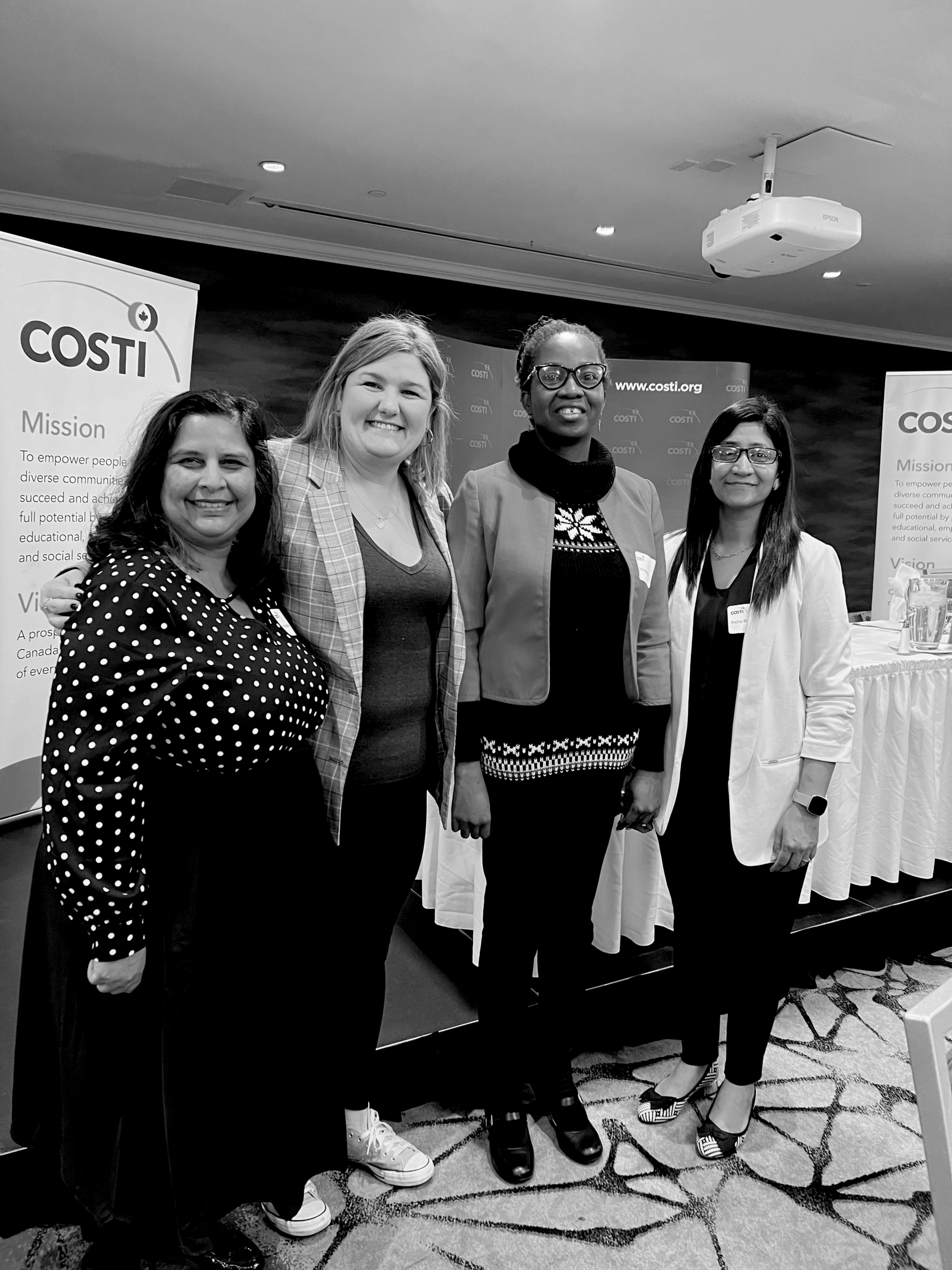 A group of four women stand side by side in front a conference stage, with COSTI standees behind them