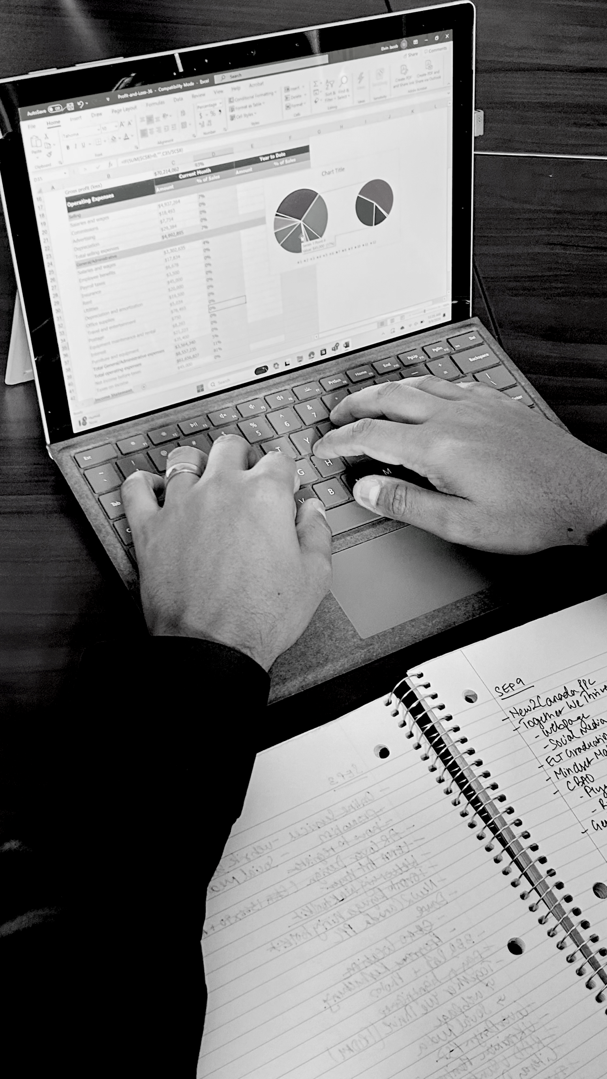 Hands typing on a laptop displaying an Excel spreadsheet with financial data and pie charts. Next to the laptop is a notebook with handwritten notes on it.