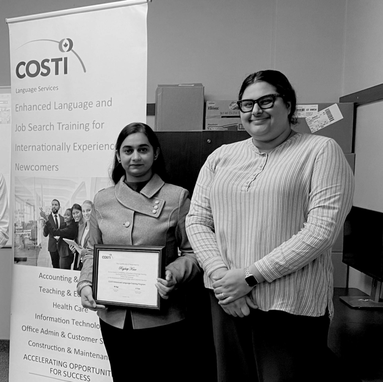 Two individuals stand together, smiling in front of a computer screen and a COSTI standee. One of them is holding a certificate.